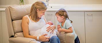Smiling mother with newborn baby in her arms and older child standing nearby looking at newborn