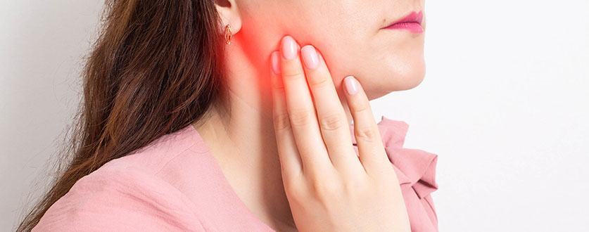 A young woman wearing a pink blouse holds her jaw, which is red and inflamed.