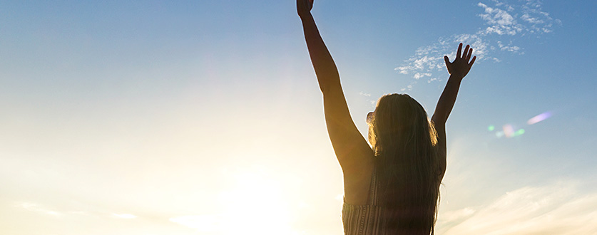 The silhouette of a young woman joyfully raising her hands in the air on a beach at sunrise.